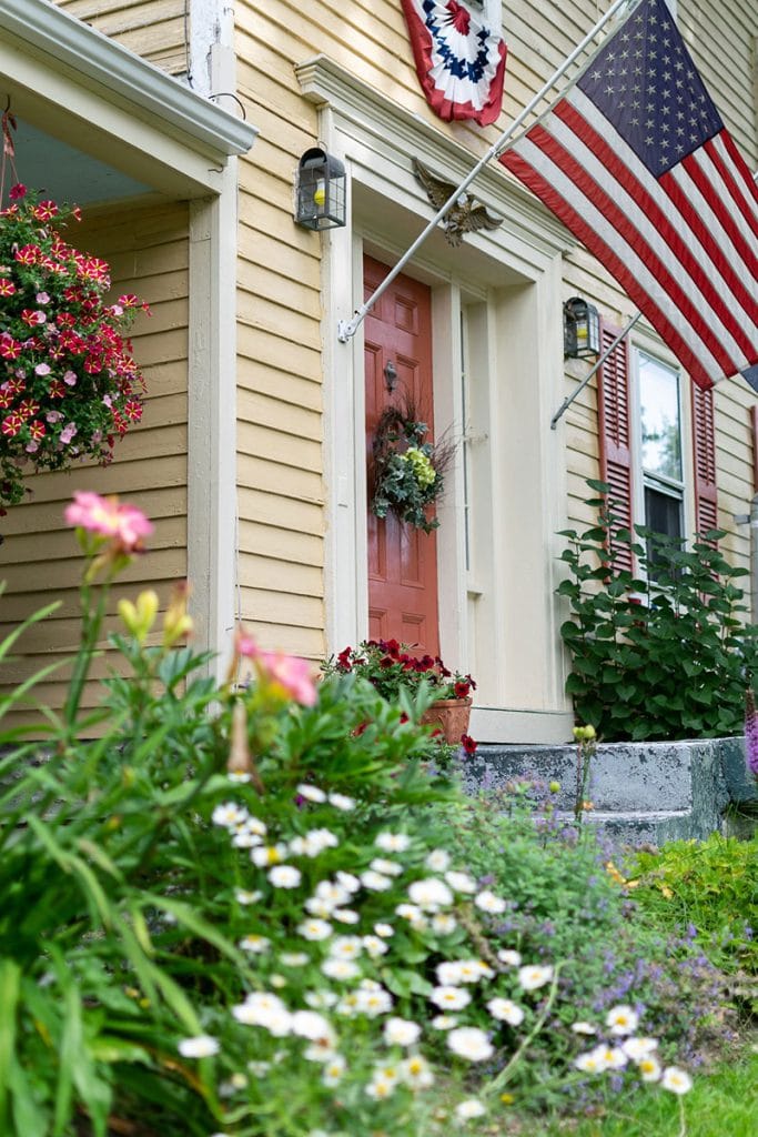 Front door of the inn with hanging flowers, summer flowers in the garden and American flag