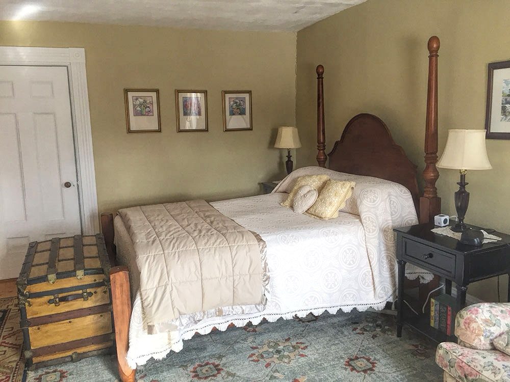 Queen bed with wooden headboard in Abigail's Suite, cream medallion bedspread and quilt on the bed, and wooden trunk at the foot of the bed. Gold walls and green oriental carpet