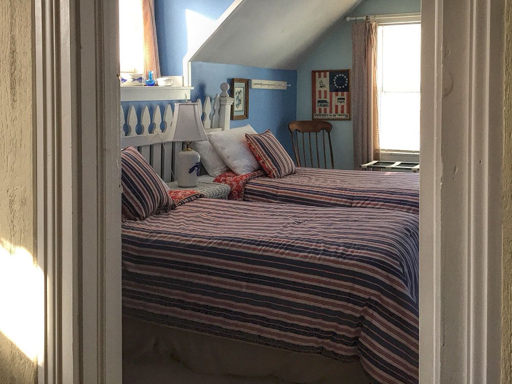Two twin beds in the second bedroom of the Ira Allen Suite with striped quilts. Blue walls in this room and a sunny window