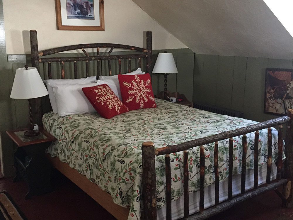 Rustic, wooden-stick queen bed in the Remember Baker Suite. Green walls and a sloping white ceiling. Wooden lamps on the night stands, and a chickadee print quilt on the bed.